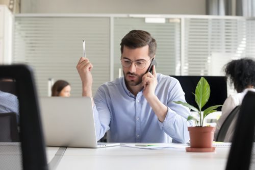 Serious confident concentrated manager employee millennial man sitting at desk in coworking space modern office with other colleagues talking by mobile phone with client solving business matters.